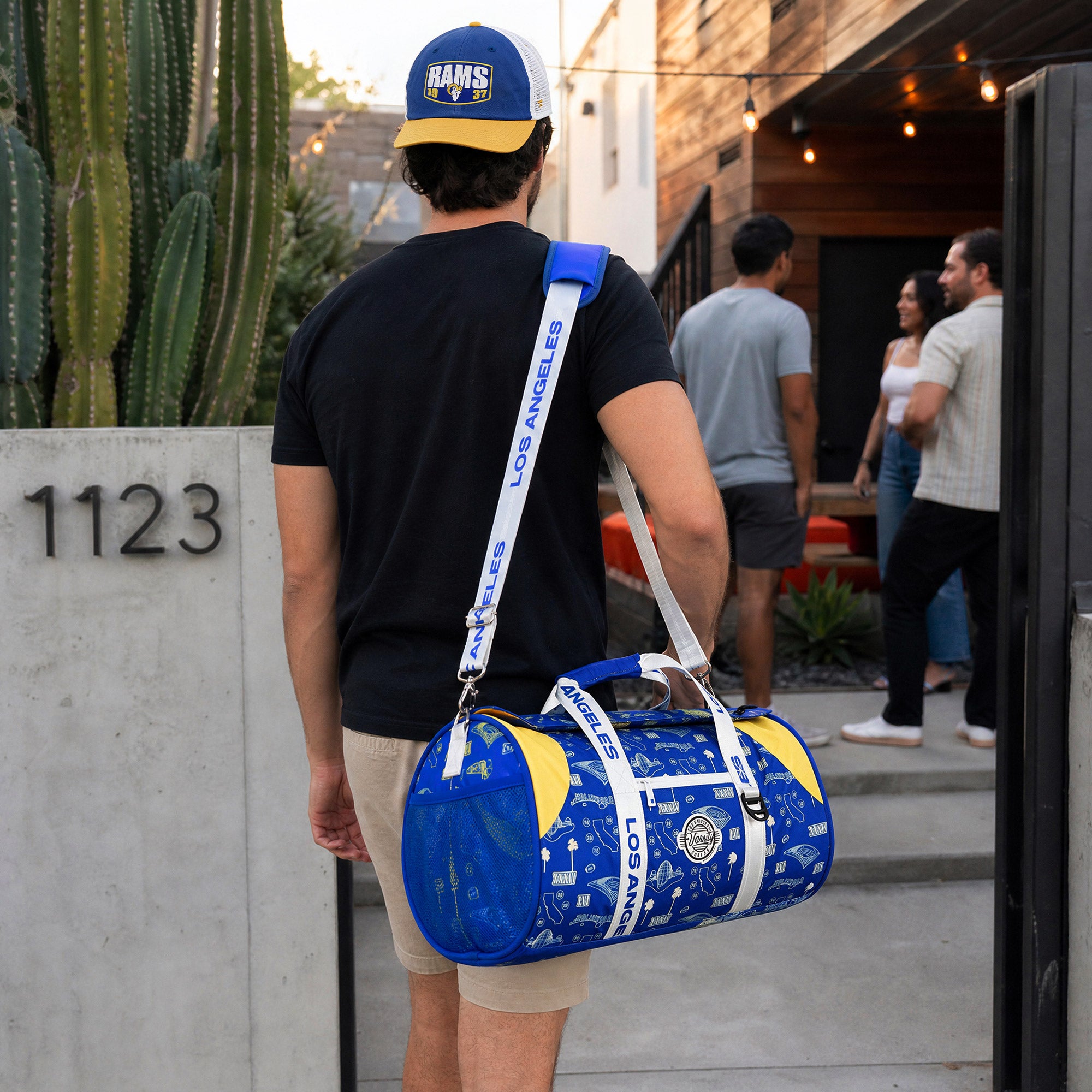 A-man-wearing-a-blue-and-yellow-Rams-cap-carries-a-blue-and-yellow-patterned-duffel-bag-with-a-white-strap-printed-with-'SEMPER-FOCO'-as-he-walks-out-of-a-house.-The-background-shows-a-patio-with-other-people-and-a-cactus.