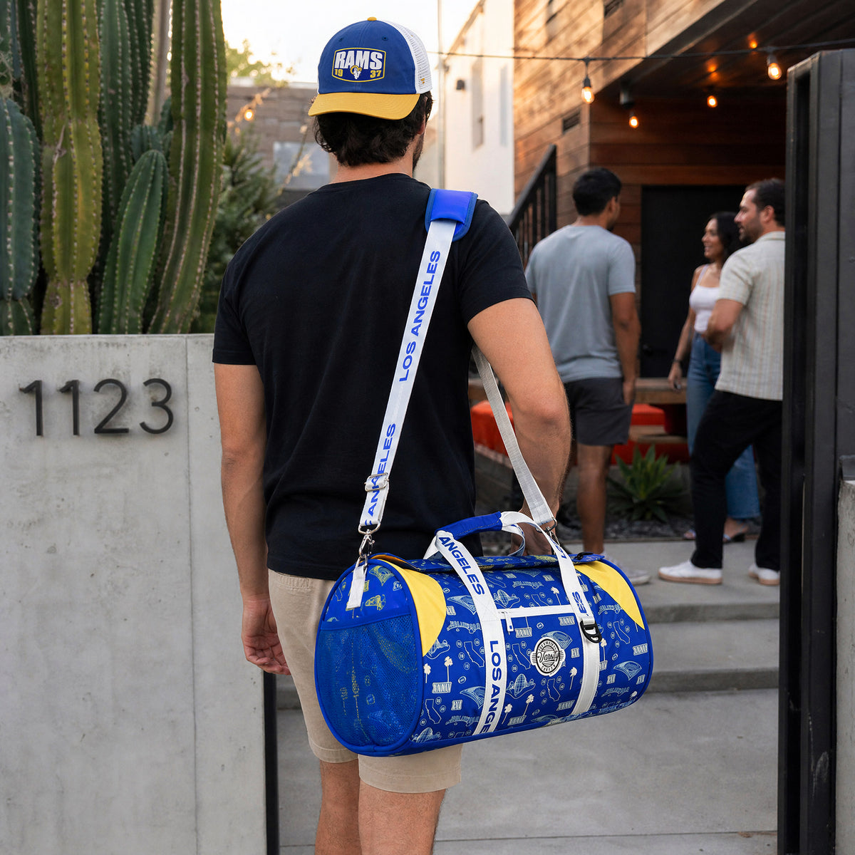 A man wearing a blue and yellow Rams cap carries a blue and yellow patterned duffel bag with a white strap printed with 'SEMPER FOCO' as he walks out of a house. The background shows a patio with other people and a cactus.