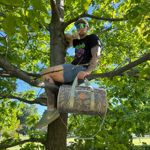 Person-sitting-in-a-tree-holding-a-duffel-bag,-surrounded-by-green-foliage.
