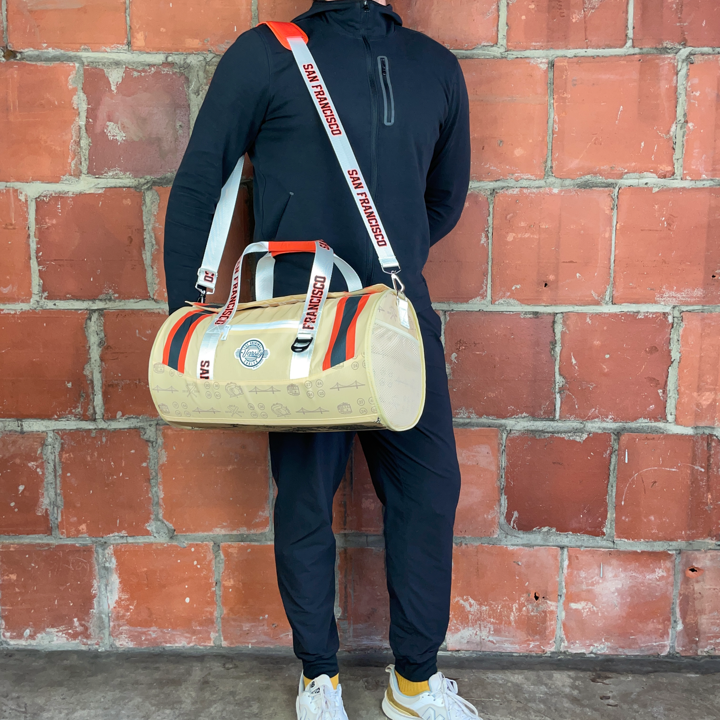 Person holding a beige duffel bag with colorful straps against a brick wall.