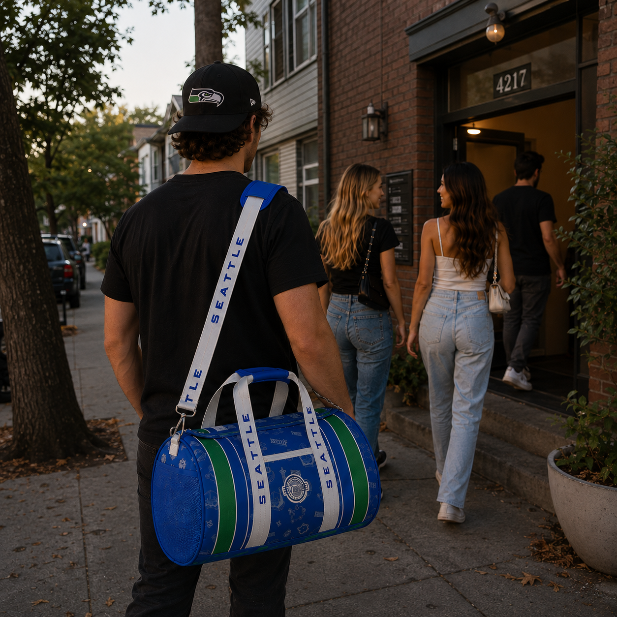 A man wearing a black Seattle Seahawks cap carries a blue Seattle Duffel bag with white and green stripes and 'SEATTLE DUFFEL' printed on the strap, walking on a city sidewalk alongside other pedestrians.